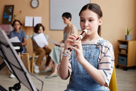 Téléchargez les photos : Écolière assise devant une partition et apprenant à jouer de la flûte en classe de musique - en image libre de droit