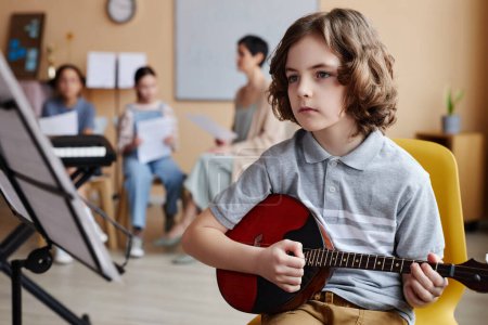 Niño sentado frente a partituras y tocando instrumentos musicales en clase de música con otros niños en el fondo