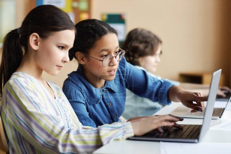 Téléchargez les photos : Écolières assis à table et travaillant sur ordinateur portable ensemble pendant les cours en classe - en image libre de droit