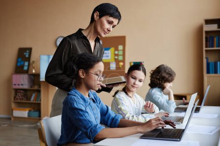Téléchargez les photos : Groupe d'écoliers utilisant des ordinateurs pendant l'étude avec un enseignant les aidant à la présentation en ligne - en image libre de droit