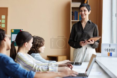 Téléchargez les photos : Enseignant utilisant la tablette PC tout en enseignant aux élèves qui sont assis à table avec des ordinateurs pendant les cours à l'école - en image libre de droit