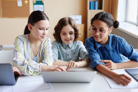 Téléchargez les photos : Des écoliers assis à table et utilisant un ordinateur portable pour faire une présentation en ligne pendant la leçon - en image libre de droit