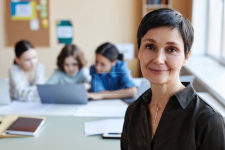 Téléchargez les photos : Portrait d'un professeur professionnel souriant à la caméra assis à la leçon avec des enfants en arrière-plan - en image libre de droit