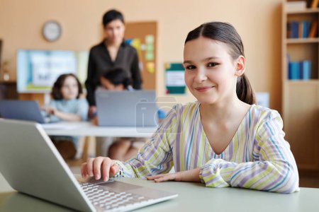 Téléchargez les photos : Portrait de l'écolière avec ordinateur portable regardant la caméra tout en étant assis à la leçon avec l'enseignant en arrière-plan - en image libre de droit