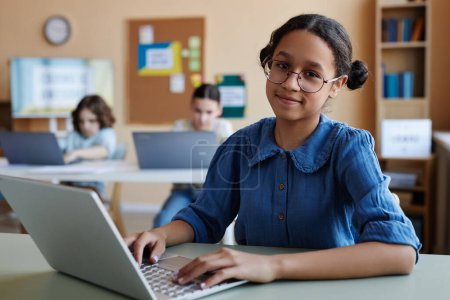 Téléchargez les photos : Portrait d'une écolière afro-américaine en lunettes souriant à la caméra assise à table et utilisant un ordinateur portable pendant les cours en classe - en image libre de droit