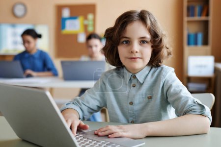 Téléchargez les photos : Portrait d'écolier regardant la caméra tout en utilisant un ordinateur portable pendant les cours en classe - en image libre de droit