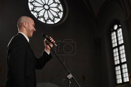 Photo for Mature pastor in black suit speaking prayer in microphone while standing in old baptist church - Royalty Free Image