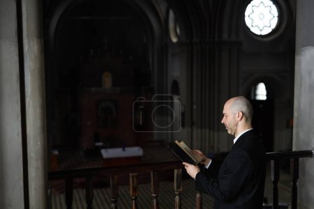Mature priest in black suit reading Bible and praying while standing in front of altar in church