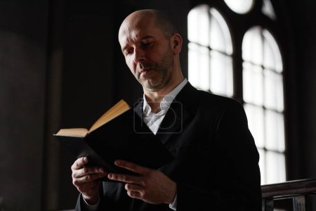 Téléchargez les photos : Concentré homme mûr en costume lecture prie dans la Bible debout dans la vieille église - en image libre de droit