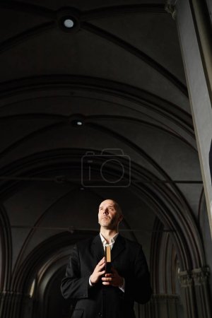 Téléchargez les photos : Image verticale de l'homme mûr avec la Bible debout dans une vieille église avec une belle architecture et en priant - en image libre de droit