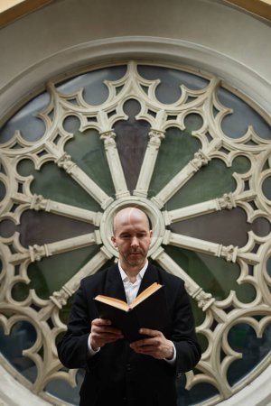 Image verticale d'un pasteur mûr sérieux en costume lisant la Bible debout contre orné en arrière-plan