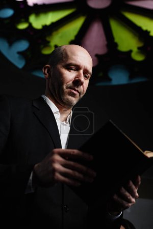 Foto de Imagen vertical del hombre maduro en traje negro leyendo la Biblia de pie en la antigua iglesia bautista - Imagen libre de derechos