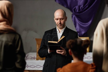 Foto de Pastor maduro en traje negro leyendo la Biblia para los creyentes de pie en la iglesia vieja - Imagen libre de derechos