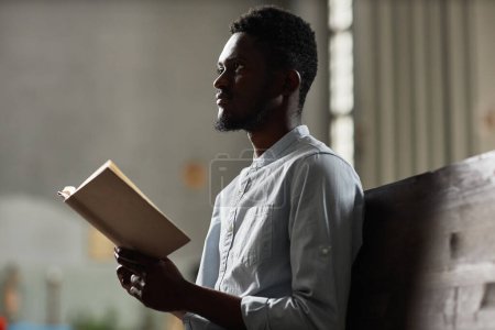 Téléchargez les photos : Afro-Américain assis sur le banc et lisant la prière du livre à l'église - en image libre de droit