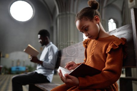 African American little girl reading Bible while sitting on bench in church with man in background