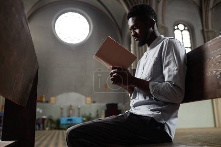 African American man sitting on bench with Bible and praying during his visit to church