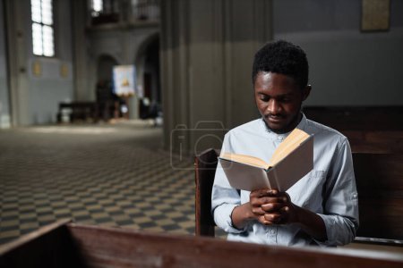 Photo for African American young man sitting on bench in church and reading prayer - Royalty Free Image