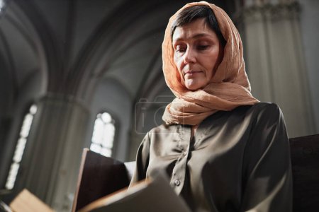 Téléchargez les photos : Femme mûre en foulard assis sur le banc dans l'église baptiste et la prière de lecture - en image libre de droit