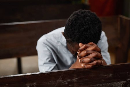 Téléchargez les photos : Jeune homme afro-américain assis sur le banc et priant dans l'église baptiste - en image libre de droit