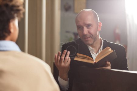 Photo for Mature pastor reading Bible and talking to boy during their meeting in church - Royalty Free Image