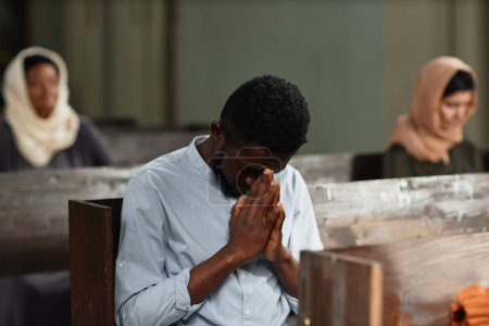 Foto de Hombre afroamericano rezando con los ojos cerrados mientras está sentado en el banco de la iglesia - Imagen libre de derechos