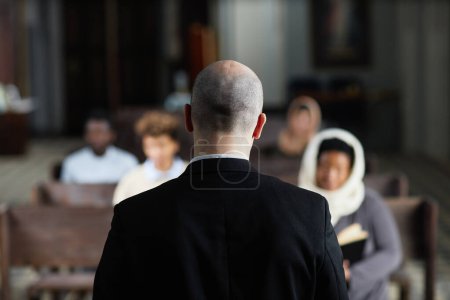 Photo for Rear view of pastor in black suit talking to believers while tehy sitting on bench in church - Royalty Free Image