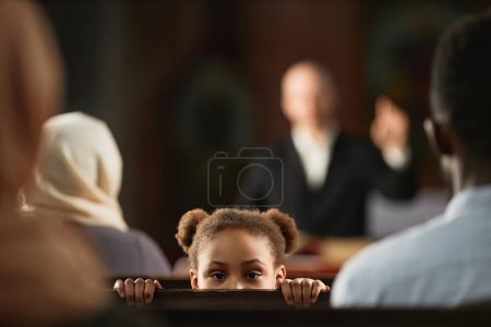 Téléchargez les photos : Portrait d'une petite fille afro-américaine regardant la caméra alors qu'elle était assise sur un banc à l'église avec d'autres personnes - en image libre de droit