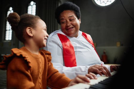 Téléchargez les photos : Femme mûre afro-américaine enseignant à une petite fille à jouer du piano pendant la représentation à l'église - en image libre de droit