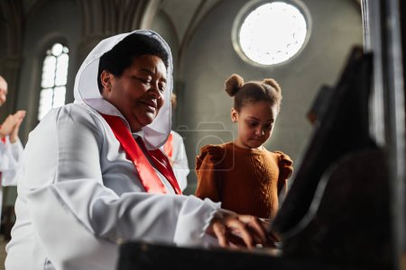 Téléchargez les photos : Femme mûre en costume blanc du choeur de l'église jouant du piano avec une petite fille - en image libre de droit