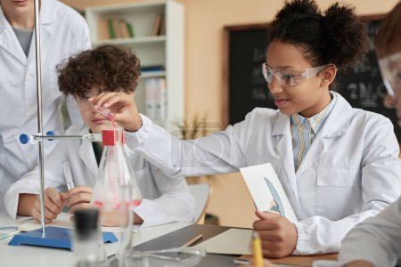 Photo for Side view portrait of smiling black schoolgirl doing science experiment in school and wearing lab coat - Royalty Free Image