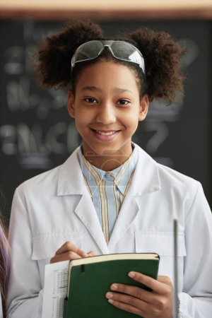 Téléchargez les photos : Portrait vertical d'une écolière noire souriante portant un manteau de laboratoire en classe de sciences et regardant une caméra tenant un cahier - en image libre de droit