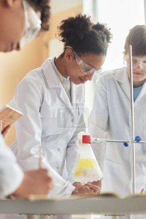 Téléchargez les photos : Portrait vertical d'une écolière noire en blouse de laboratoire en classe de sciences et faisant des expériences - en image libre de droit