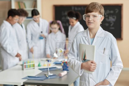 Photo for Waist up portrait of teen schoolboy wearing lab coat in science class and looking at camera with group of children in background, copy space - Royalty Free Image