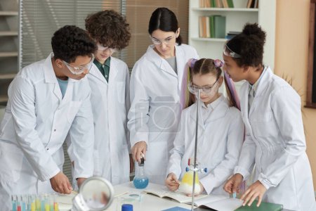 Téléchargez les photos : Groupe diversifié de jeunes adolescents portant des blouses de laboratoire faisant des expériences scientifiques à l'école avec l'aide d'enseignants - en image libre de droit