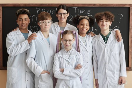 Téléchargez les photos : Groupe diversifié d'enfants portant des blouses de laboratoire posant avec le professeur en classe de sciences à l'école et souriant à la caméra - en image libre de droit