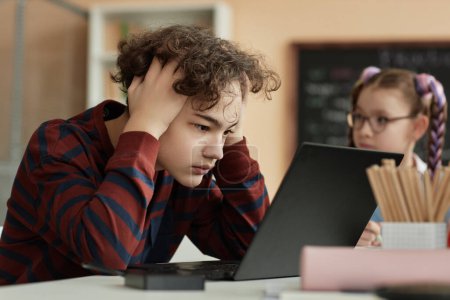 Téléchargez les photos : Portrait latéral d'un écolier adolescent en détresse utilisant un ordinateur à l'école et passant un examen en ligne - en image libre de droit