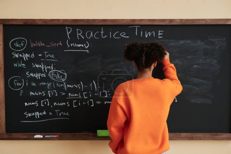 Téléchargez les photos : Vue arrière de l'adolescente noire écrivant sur le tableau noir dans la classe de l'école, espace de copie - en image libre de droit