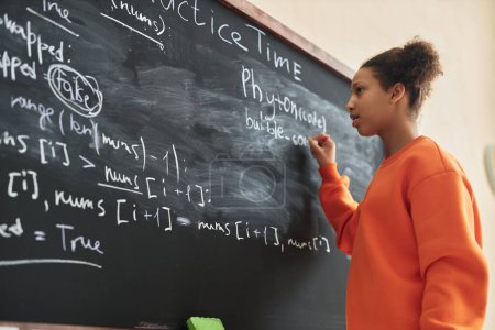 Téléchargez les photos : Vue latérale portrait d'une adolescente noire écrivant sur un tableau noir dans une classe d'école, espace de copie - en image libre de droit