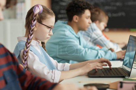 Téléchargez les photos : Vue latérale des écoliers utilisant des ordinateurs en rangée et passant un examen en ligne - en image libre de droit