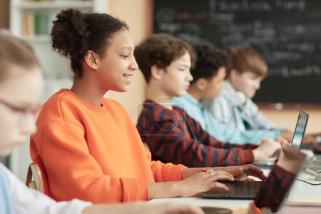 Téléchargez les photos : Vue latérale à une adolescente noire utilisant des ordinateurs en rangée dans la salle de classe de l'école et prenant examen en ligne - en image libre de droit