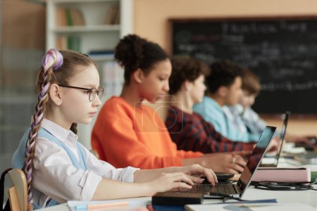 Téléchargez les photos : Vue latérale à l'école mignonne en utilisant l'ordinateur en rangée dans la salle de classe de l'école et de prendre examen en ligne - en image libre de droit