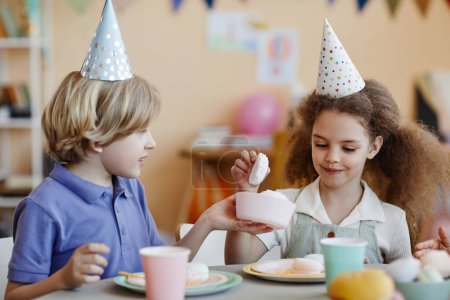 Foto de Retrato de dos niños felices usando sombreros de fiesta y disfrutando de dulces en la mesa - Imagen libre de derechos