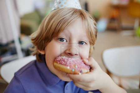 Gros plan portrait de petit garçon blond mangeant un gros beignet pendant la fête d'anniversaire et regardant la caméra