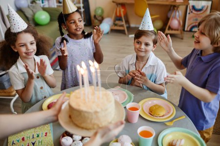 Foto de Vista de ángulo alto en un grupo diverso de niños felices aplaudiendo mientras esperan pastel con velas durante la fiesta de cumpleaños - Imagen libre de derechos