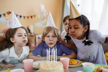 Diverse group of happy children blowing candles on cake together during birthday party