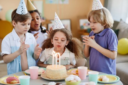 Portrait de mignonne petite fille soufflant des bougies sur le gâteau pendant la fête d'anniversaire avec des amis.