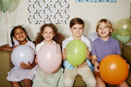 Foto de Diverso grupo de niños sosteniendo globos y sonriendo a la cámara durante la fiesta para niños disparados con flash - Imagen libre de derechos