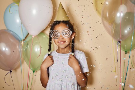 Portrait taille haute de petite fille noire avec des ballons portant des lunettes funky à la fête et des paillettes tombant