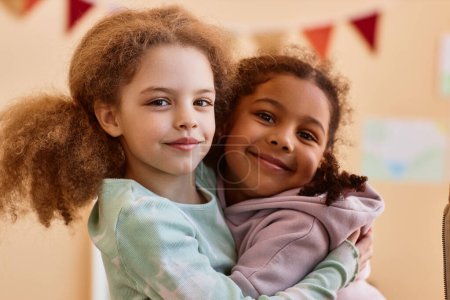 Photo for Portrait of two cute little girls embracing and smiling at camera at home - Royalty Free Image