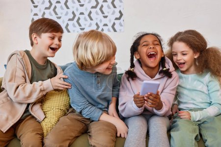 Groupe de petits enfants joyeux riant émotionnellement et utilisant un smartphone à l'intérieur